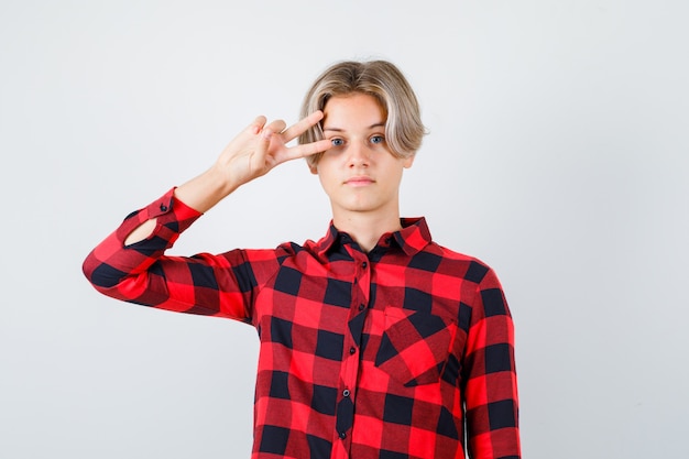 Giovane ragazzo adolescente in camicia a quadri che mostra il segno V vicino all'occhio e sembra fiducioso, vista frontale.