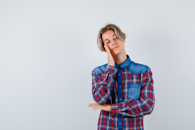 Giovane ragazzo adolescente in camicia a quadri che si appoggia la guancia a portata di mano e sembra assonnato, vista frontale.