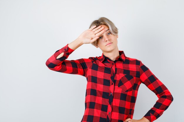 Giovane ragazzo adolescente in camicia a quadri con la mano sulla fronte e guardando triste, vista frontale.