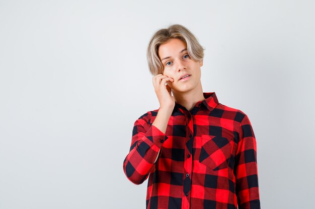 Giovane ragazzo adolescente in camicia a quadri con la mano sulla guancia e guardando fiducioso, vista frontale.