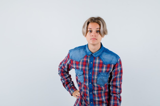 Giovane ragazzo adolescente in camicia a quadri tenendo la mano sulla vita e guardando perplesso, vista frontale.