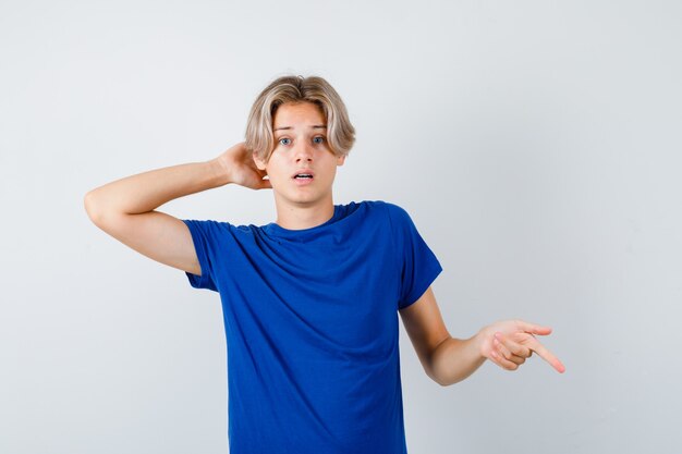 Giovane ragazzo adolescente in maglietta blu con la mano dietro la testa, che punta verso il basso e sembra confuso, vista frontale.