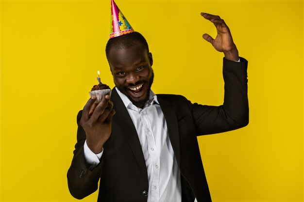 Giovane ragazzo afroamericano sorridente in abito nero e cappello compleanno con candela accesa