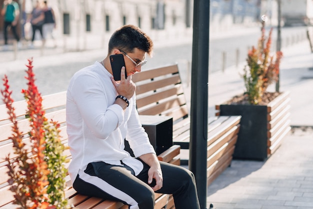 Giovane ragazzo alla moda in camicia con il telefono sul banco in giornata di sole caldo all'aperto