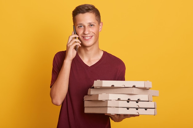 Giovane ragazzo allegro in piedi isolato su giallo in studio, tenendo le scatole di cartone con pizza e smartphone in mano, conversando, parlando al telefono, guardando direttamente la fotocamera.