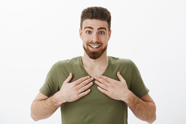 Giovane ragazzo barbuto felice grato e stupito sorpreso in maglietta verde oliva che alza le sopracciglia sorridendo stupito ed eccitato ampiamente stordito con un regalo inaspettato che tiene i palmi sul petto