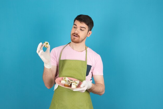 Giovane ragazzo barbuto guardando i rotoli di torta fatta in casa su un blu.