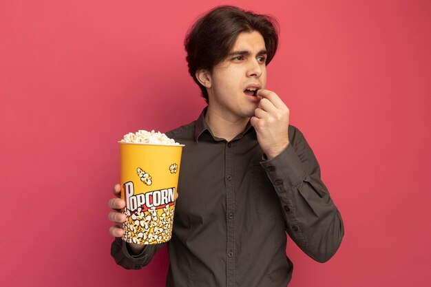 Giovane ragazzo bello che guarda al lato che indossa la maglietta nera che tiene e che prova il secchio di popcorn isolato sulla parete rosa