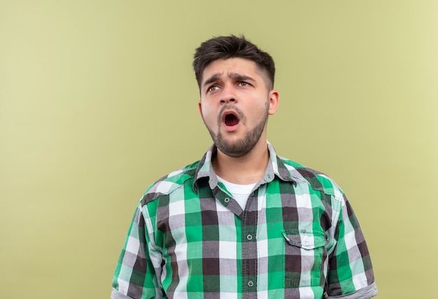 Giovane ragazzo bello che indossa la camicia a scacchi che sbadiglia in piedi stanco sopra la parete cachi