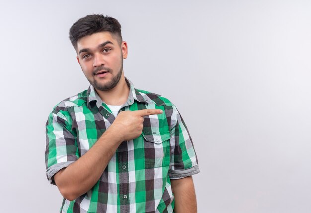 Giovane ragazzo bello che indossa la camicia a scacchi che sembra sorpreso che punta a sinistra con l'indice destro in piedi sopra il muro bianco