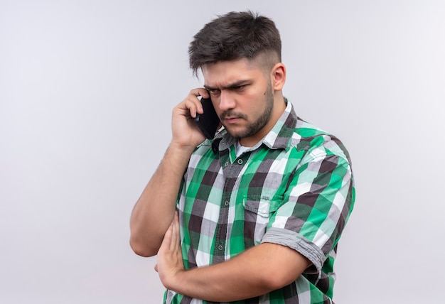 Giovane ragazzo bello che indossa la camicia a scacchi parlando con rabbia al telefono in piedi sopra il muro bianco