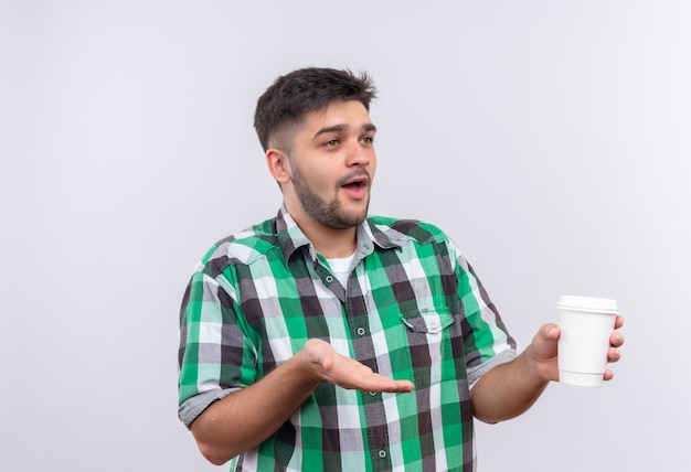 Giovane ragazzo bello che indossa la camicia a scacchi sorpreso a parlare con qualcuno che tiene la tazza di caffè di plastica in piedi sopra il muro bianco