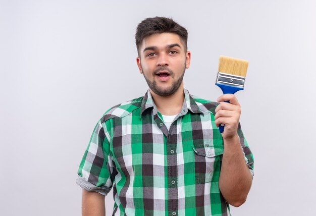 Giovane ragazzo bello che indossa la camicia a scacchi tenendo il pennello in piedi sul muro bianco