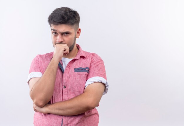 Giovane ragazzo bello che indossa la camicia di polo rosa che osserva in piedi sopra il muro bianco