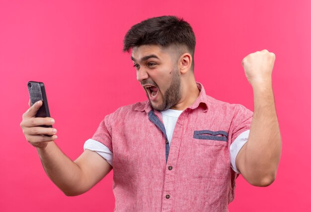 Giovane ragazzo bello che indossa la camicia di polo rosa che tiene il telefono, facendo segno di sì con il pugno alzato in piedi sopra il muro rosa