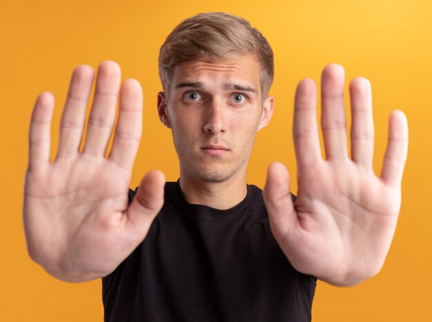 Giovane ragazzo bello che indossa la camicia nera che mostra il gesto di arresto isolato sulla parete gialla