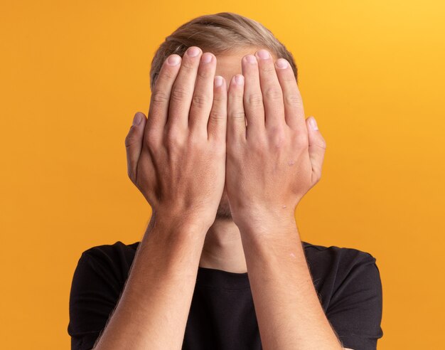 Giovane ragazzo bello che indossa la camicia nera ha coperto il viso con le mani isolate sul muro giallo