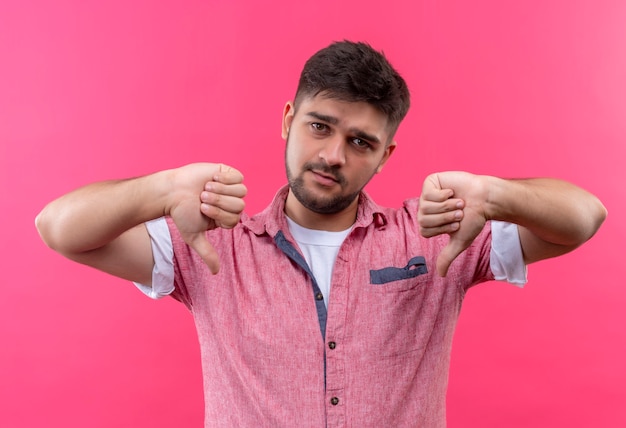 Giovane ragazzo bello che indossa la maglietta di polo rosa che fa i pollici infelici giù in piedi sopra il muro rosa