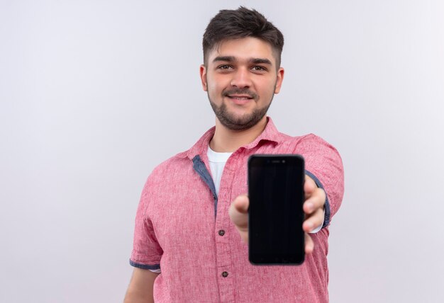 Giovane ragazzo bello che indossa la maglietta di polo rosa che guarda felicemente che mostra il telefono che sta sopra il muro bianco