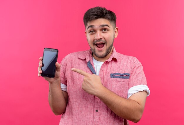 Giovane ragazzo bello che indossa la maglietta polo rosa che punta felicemente al telefono con l'indice in piedi sopra il muro rosa