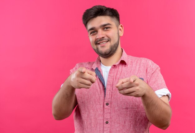 Giovane ragazzo bello che indossa la maglietta polo rosa che punta felicemente alla telecamera con gli indici che fanno hey si firma in piedi sopra il muro rosa