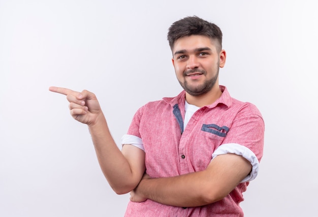 Giovane ragazzo bello che indossa la maglietta polo rosa che sorride coridally che indica a destra con l'indice che sta sopra il muro bianco