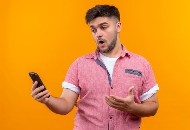 Giovane ragazzo bello che indossa la maglietta polo rosa guardando il telefono, in piedi sul muro arancione