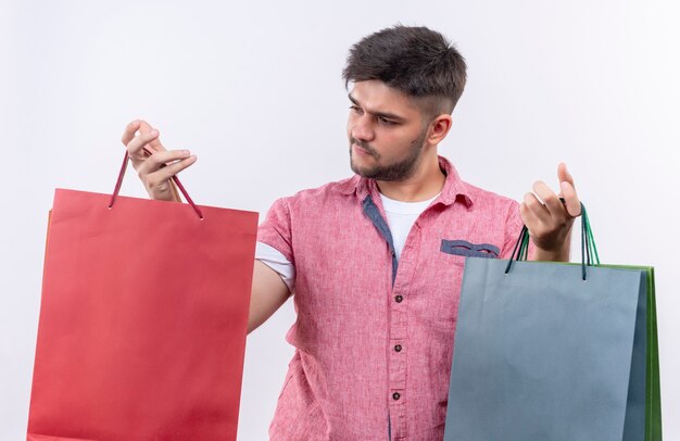 Giovane ragazzo bello che indossa la maglietta polo rosa indeciso guardando la borsa rossa in piedi sopra il muro bianco