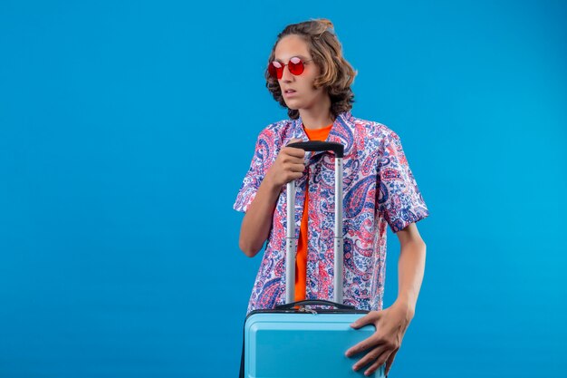 Giovane ragazzo bello che indossa occhiali da sole rossi che tengono la valigia di viaggio che sembra confuso in piedi su sfondo blu