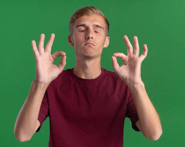 Giovane ragazzo bello con gli occhi chiusi che indossa la camicia rossa che mostra gesto di meditazione isolato sulla parete verde