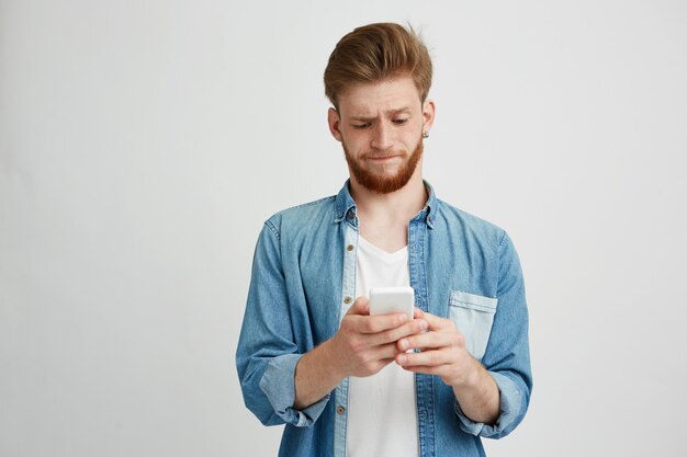 Giovane ragazzo bello dispiaciuto che esamina lo schermo del telefono che passa in rassegna web.