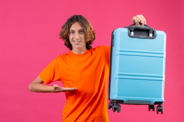 Giovane ragazzo bello in valigia arancione di viaggio della holding della maglietta che indica con il braccio della mano che sorride con la condizione felice del fronte