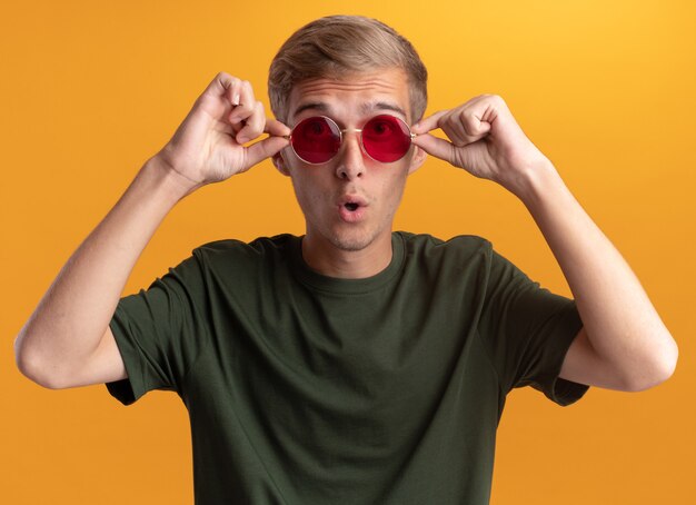 Giovane ragazzo bello sorpreso che indossa camicia verde e occhiali isolati sulla parete gialla