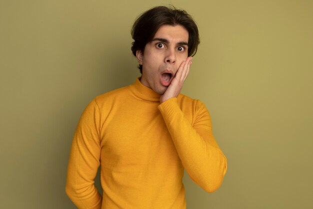 Giovane ragazzo bello sorpreso che indossa maglione dolcevita giallo che mette la mano sulla guancia isolata sulla parete verde oliva