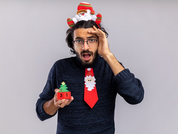 Giovane ragazzo bello spaventato che indossa il legame di natale con il cerchio dei capelli che tiene il giocattolo di natale che mette la mano sulla fronte isolata su fondo bianco