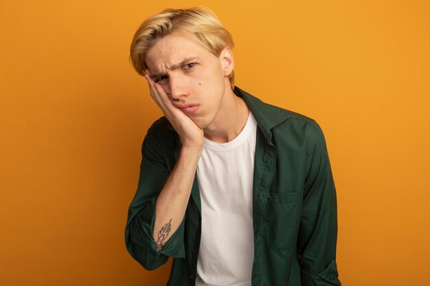 Giovane ragazzo biondo dispiaciuto che indossa la maglietta verde che mette la mano sulla guancia