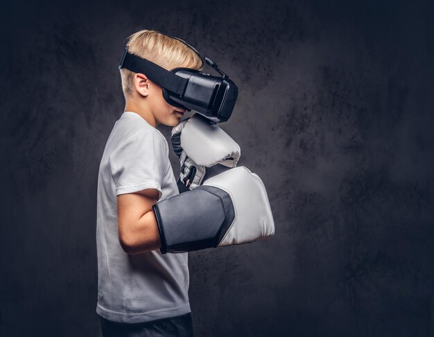 Giovane ragazzo boxer con capelli biondi vestito con una t-shirt bianca che indossa occhiali per realtà visiva e guantoni da boxe, allenamento in studio. Isolato su uno sfondo scuro con texture.