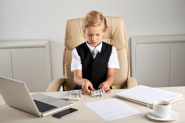 Giovane ragazzo caucasico che si siede nella sedia esecutiva in ufficio e che conta i dollari sullo scrittorio