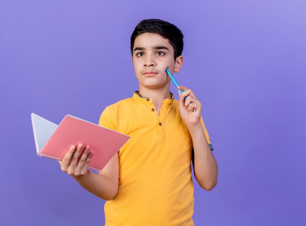 Giovane ragazzo caucasico premuroso che tiene blocco note toccando la guancia con la matita guardando il lato isolato sulla parete viola con lo spazio della copia