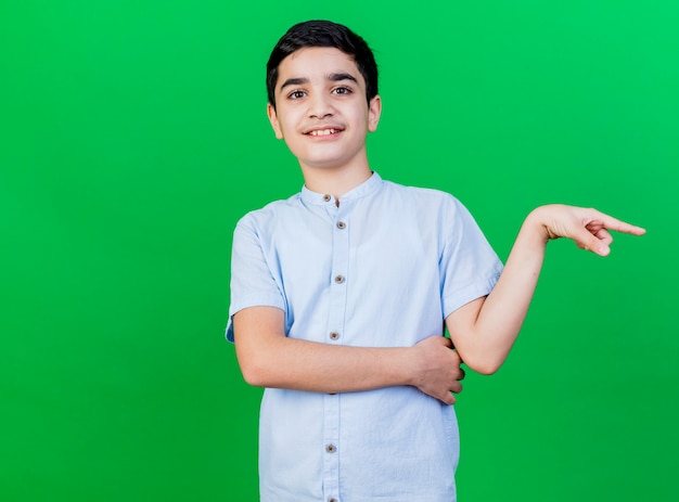 Giovane ragazzo caucasico sorridente che indica al lato isolato sulla parete verde con lo spazio della copia