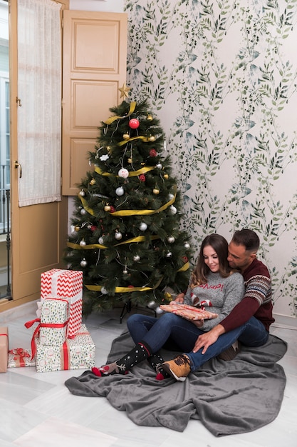 Giovane ragazzo che abbraccia da retro signora allegra con scatola regalo seduto vicino albero di Natale