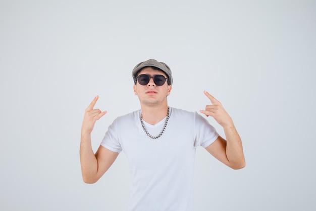 Giovane ragazzo che mostra gesto di roccia in t-shirt, cappello e guardando sicuro di sé, vista frontale.