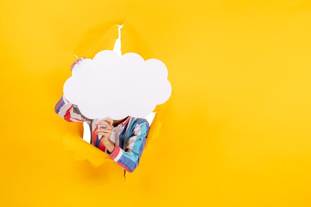 Giovane ragazzo che tiene carta bianca a forma di nuvola davanti al suo viso e posa per la macchina fotografica in un buco strappato e uno sfondo libero in carta gialla