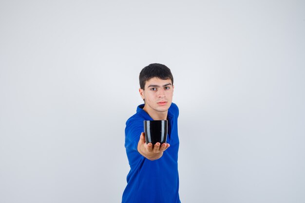 Giovane ragazzo che tiene la tazza, dandola a qualcuno in maglietta blu e guardando serio, vista frontale.