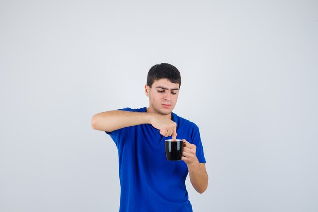 Giovane ragazzo che tiene la tazza vicino al mento, mettendoci la mano in maglietta blu e guardando curioso, vista frontale.