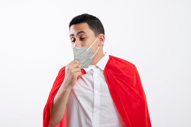 Giovane ragazzo del supereroe in mantello rosso che indossa la maschera protettiva afferrando la maschera cercando di toglierlo con gli occhi chiusi isolati su priorità bassa bianca