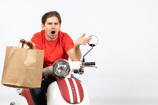 Giovane ragazzo di consegna scioccato in uniforme rossa che si siede sul motorino che tiene il sacchetto di carta sul muro bianco