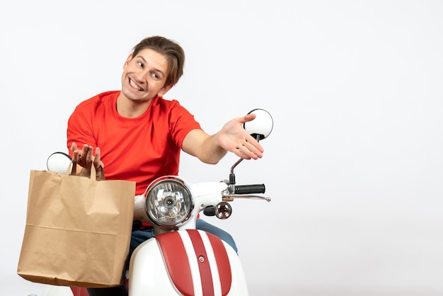 Giovane ragazzo di consegna sorridente in uniforme rossa che si siede sul motorino che tiene il sacchetto di carta e dà il benvenuto al muro bianco