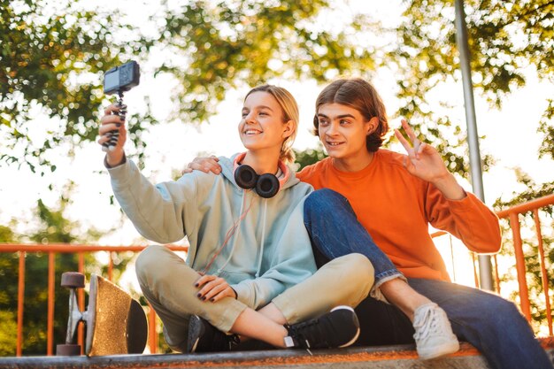 Giovane ragazzo e ragazza sorridenti skater con le cuffie che registrano felicemente un nuovo video insieme allo skatepark moderno