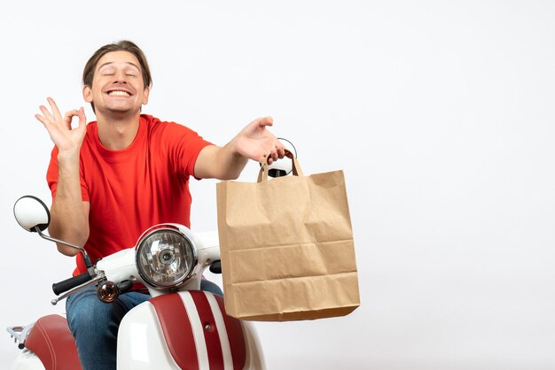 Giovane ragazzo emotivo sorridente del corriere in uniforme rossa che si siede sul motorino che dà il sacchetto di carta che fa gesto perfetto sulla parete bianca